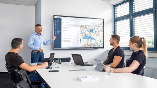 Ein Flottenmanager belehrt ein Team aus zwei Männern und einer Frau in einem hellen, modernen Besprechungsraum. Dabei deutet er auf einen großen Bildschirm und sein Team hört ihm aufmerksam zu.