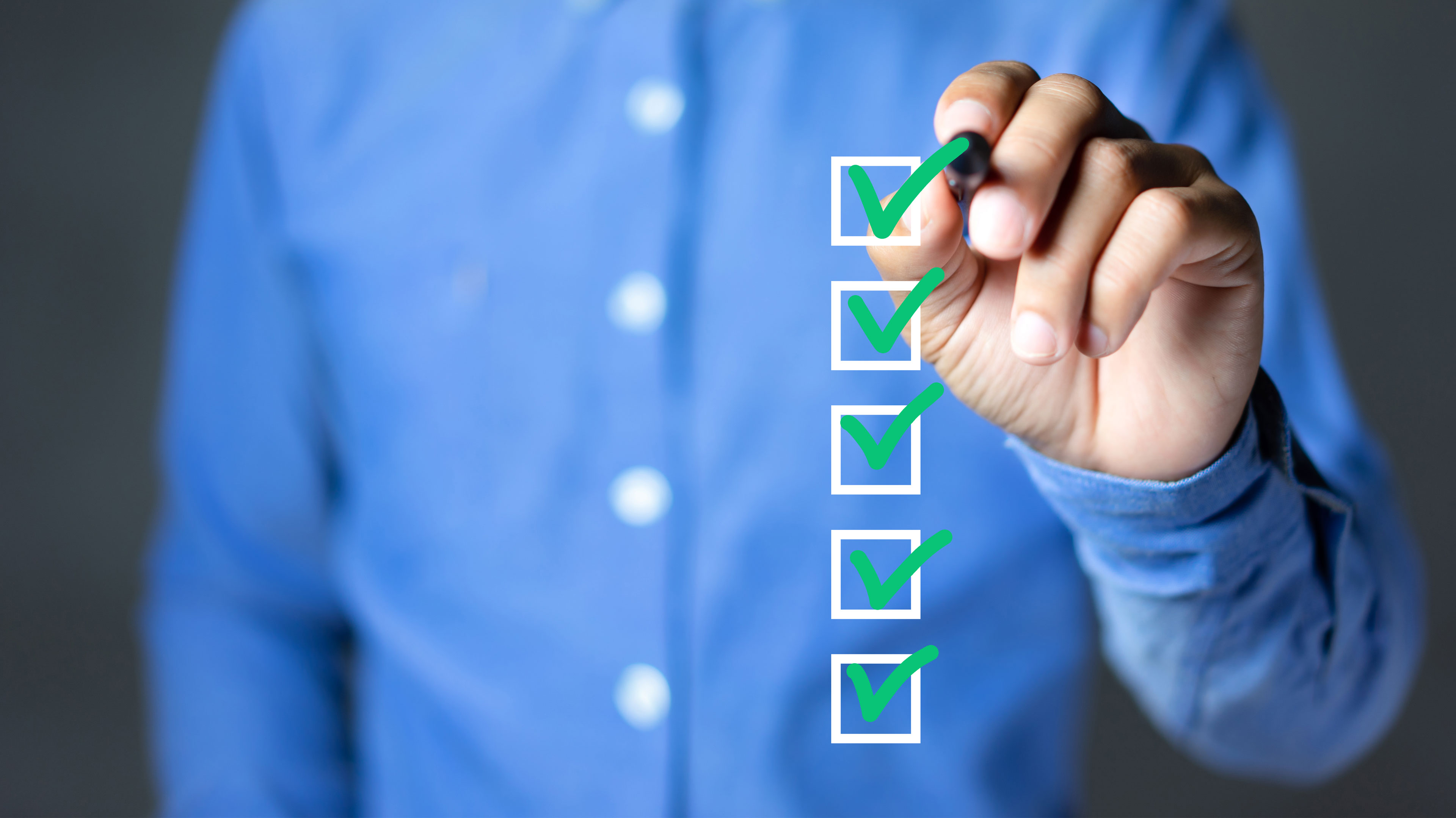 Man sieht den Oberkörper eines Mannes, der ein blaues Hemd trägt und einen Stift in der Hand hält. Damit hakt er weiße Felder an, die in einer Fotomontage über das Bild gelegt sind. Alle Felder sind mit einem grünen Haken versehen.