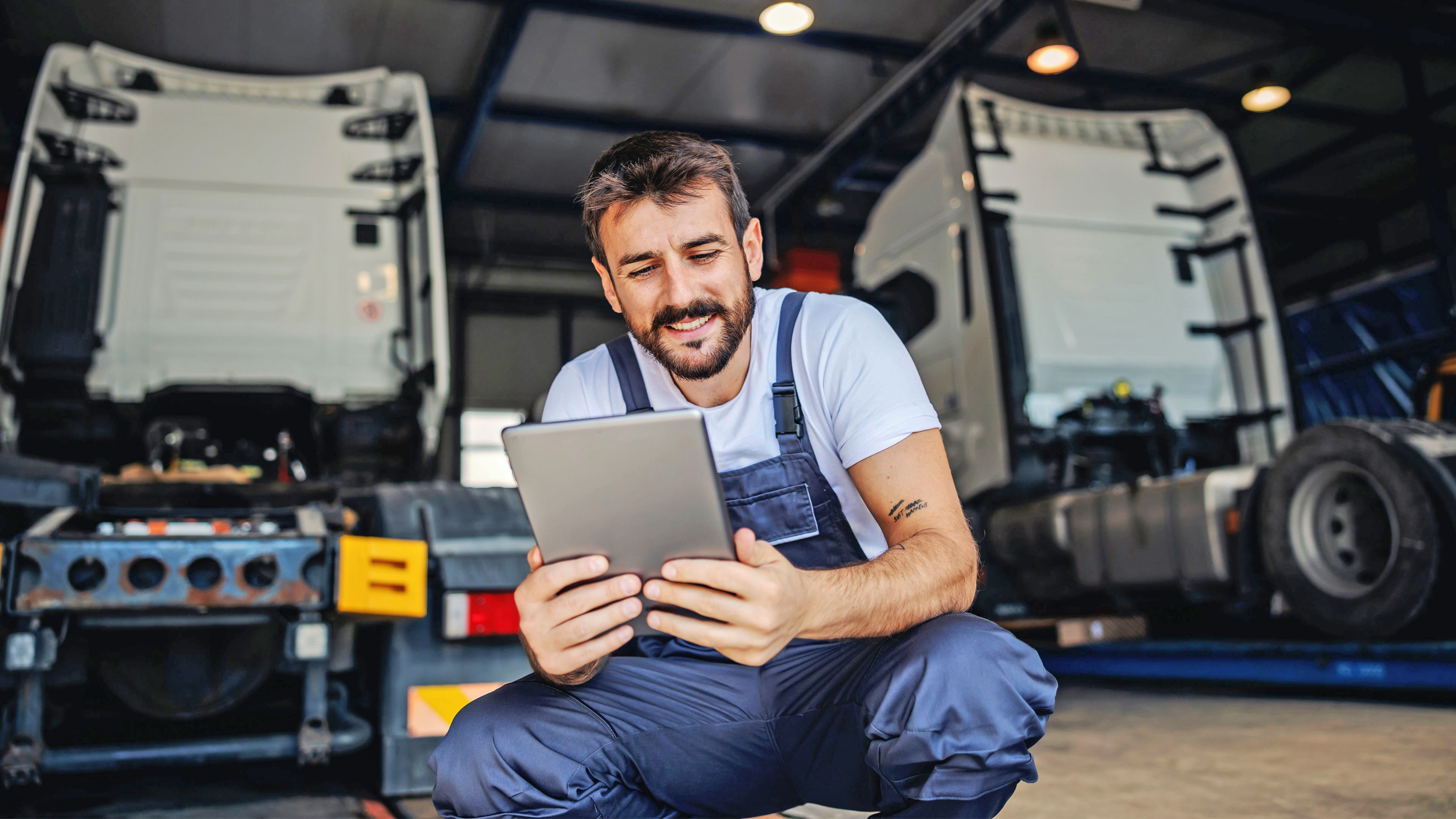 Man sieht im Vordergrund einen Mann in blauer Arbeitskleidung, der ein Tablet in der Hand hält und interessiert darauf schaut. Er ist in der Hocke, sodass man hinter ihm die Umgebung seiner Werkstatt und zwei Lkw erkennen kann.